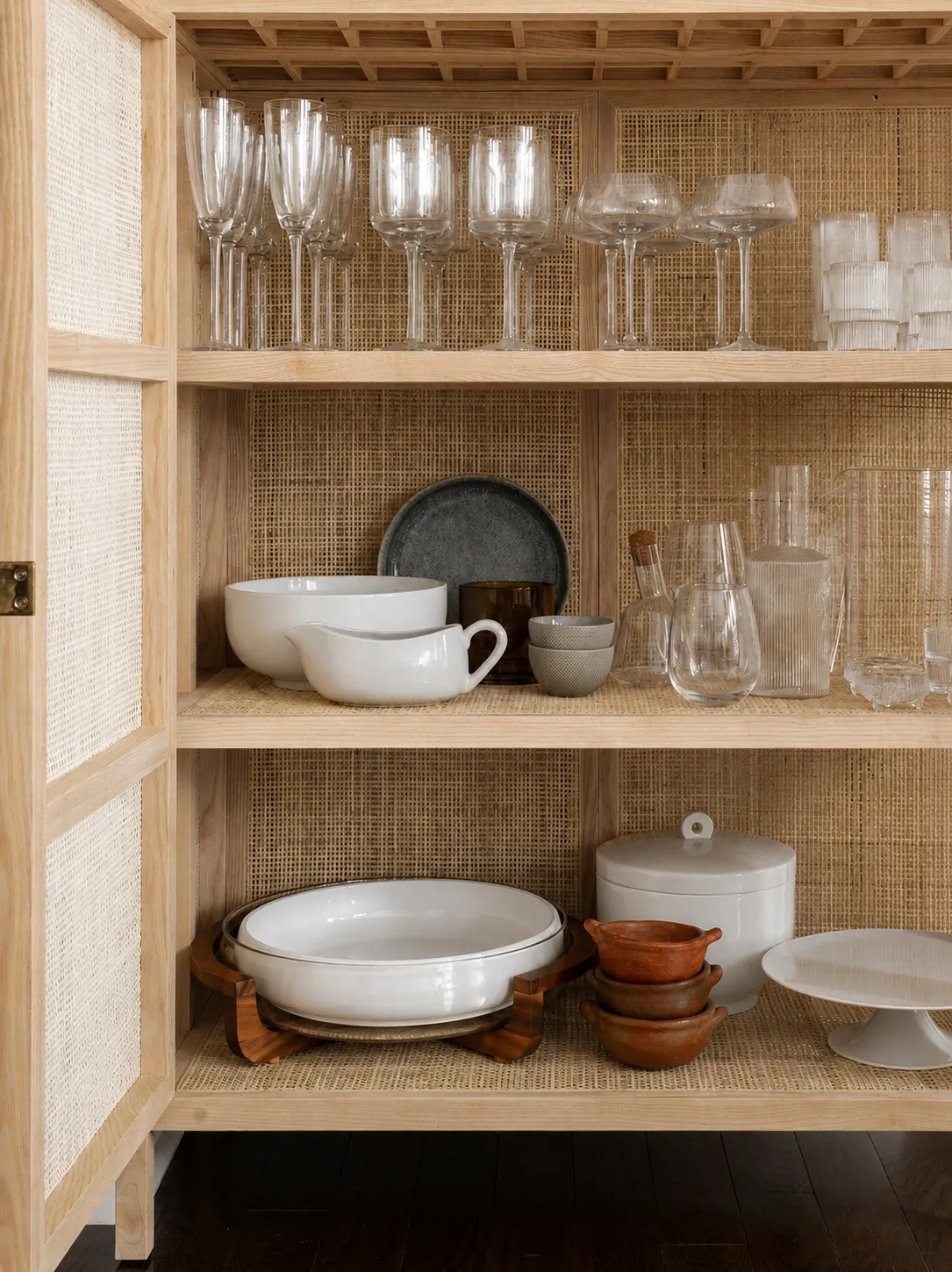A wooden cabinet with woven panels displays glassware, plates, bowls, pitchers, and carafes on three shelves. The top shelf holds glasses, the middle has dishes and cups, and the bottom features serving bowls and wooden trays.