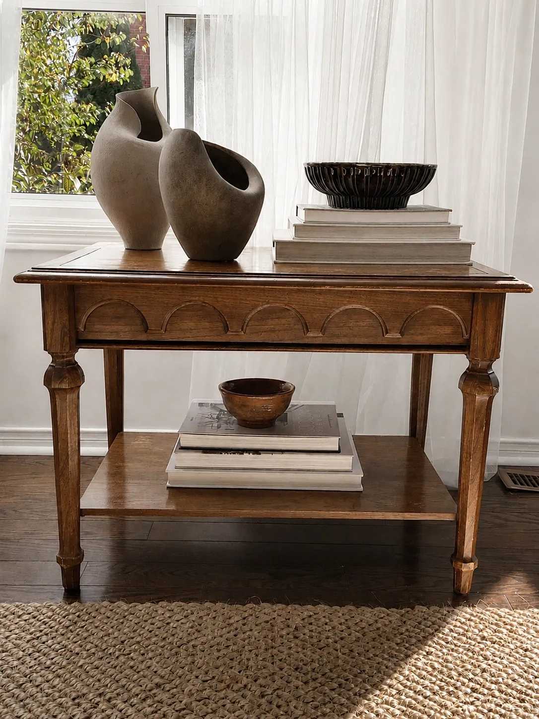 A wooden console table with decorative vases, stacked books, and a bowl sits on a woven rug by a bright window, with leafy greenery visible outside.