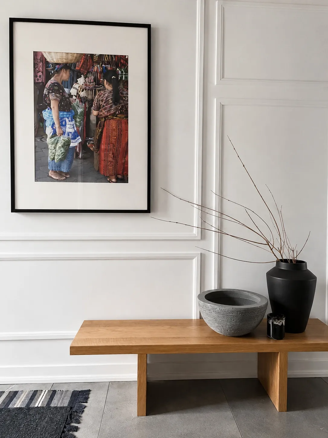 A minimalist entryway with a wooden bench, a black vase with bare branches, a gray bowl, and a candle. Above the bench hangs a framed photo of people in colorful clothing at a market.