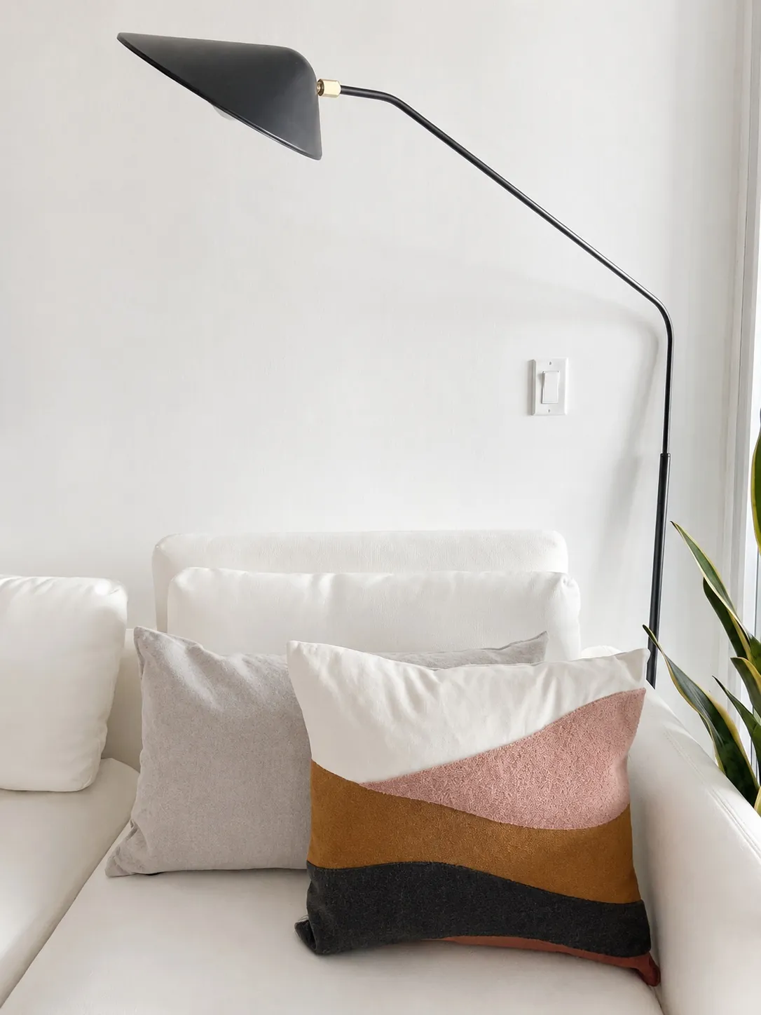 A modern living room corner with a white sofa, two decorative pillows (one plain gray, one with abstract color blocks), a black floor lamp with an angled arm, and part of a green leafy plant.