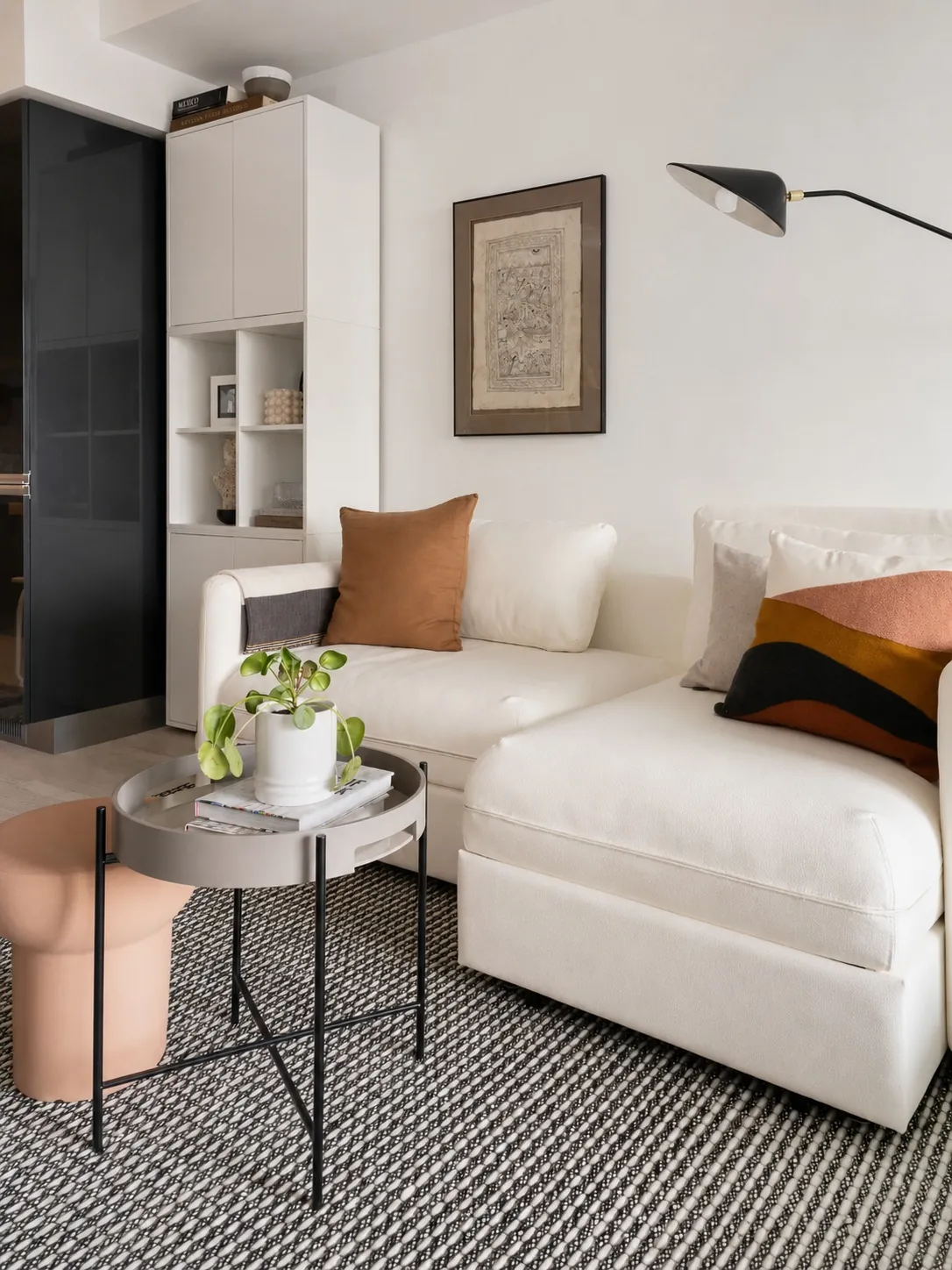A modern living room with a white sectional sofa, orange and brown accent pillows, a round gray coffee table with a potted plant, a pink ottoman, striped rug, wall art, and a black floor lamp.