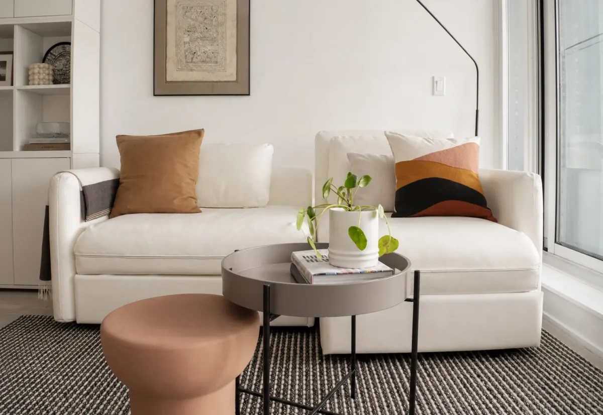 A modern living room with two white sofas, earth-tone cushions, a round gray coffee table with a potted plant and books, a brown ottoman, a wall-mounted art piece, and large windows letting in natural light.