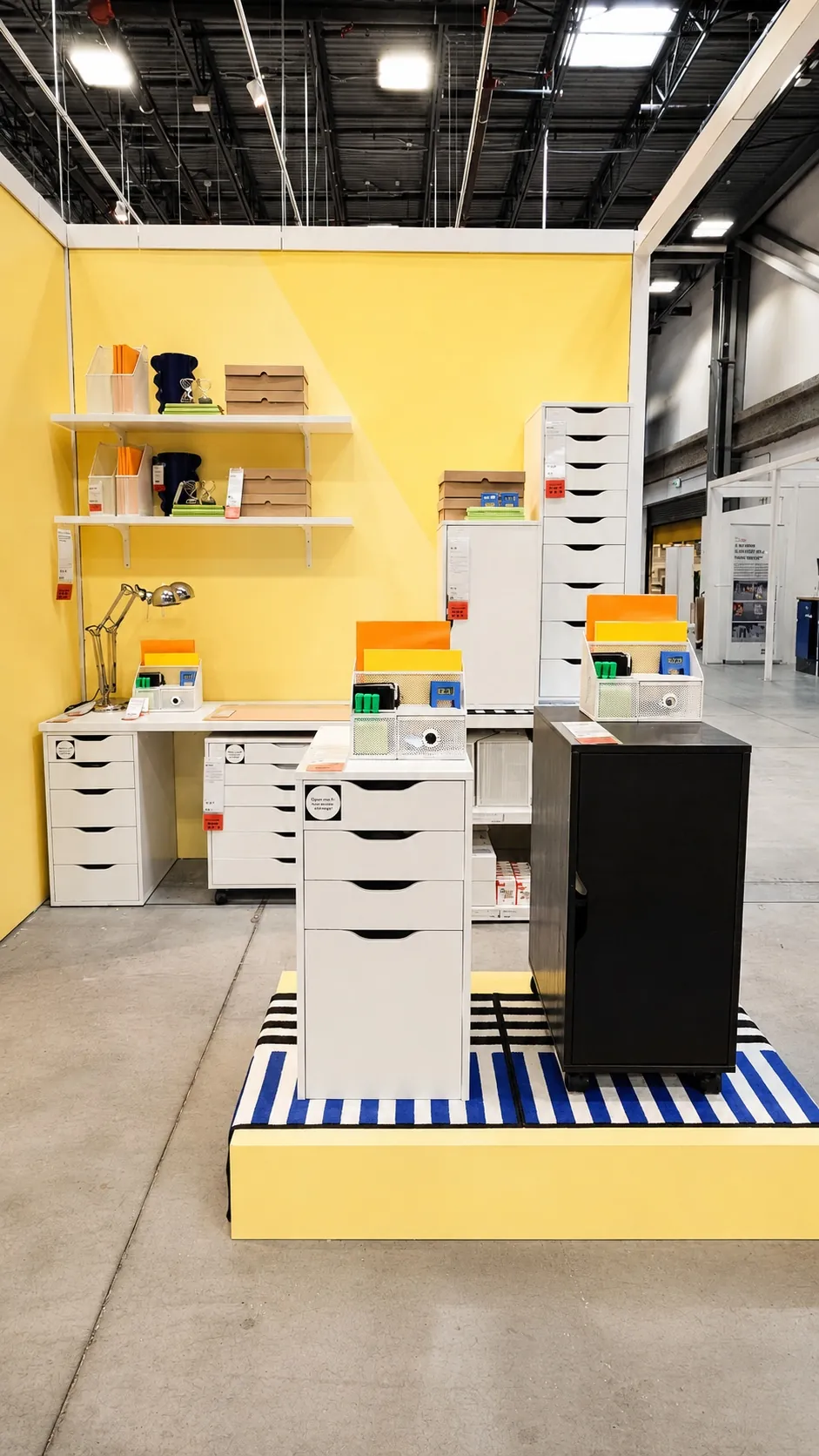 A showroom display features white filing cabinets and desks with colorful file organizers, books, and a lamp, all set against a yellow wall and blue-striped platform in a spacious warehouse setting.