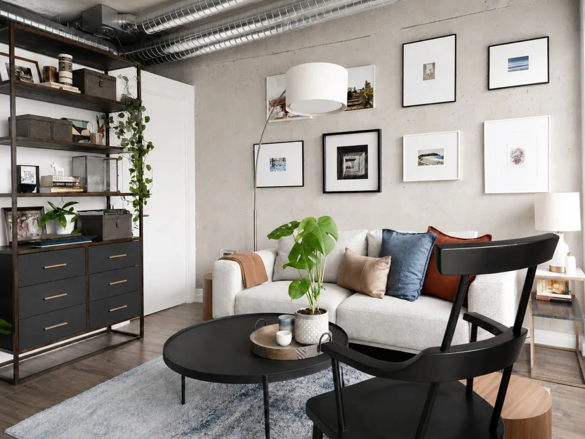 A modern living room with a light-gray sofa, colorful cushions, a black chair, round coffee tables, a potted plant, shelves with decor, framed art on a concrete wall, and exposed industrial ductwork on the ceiling.