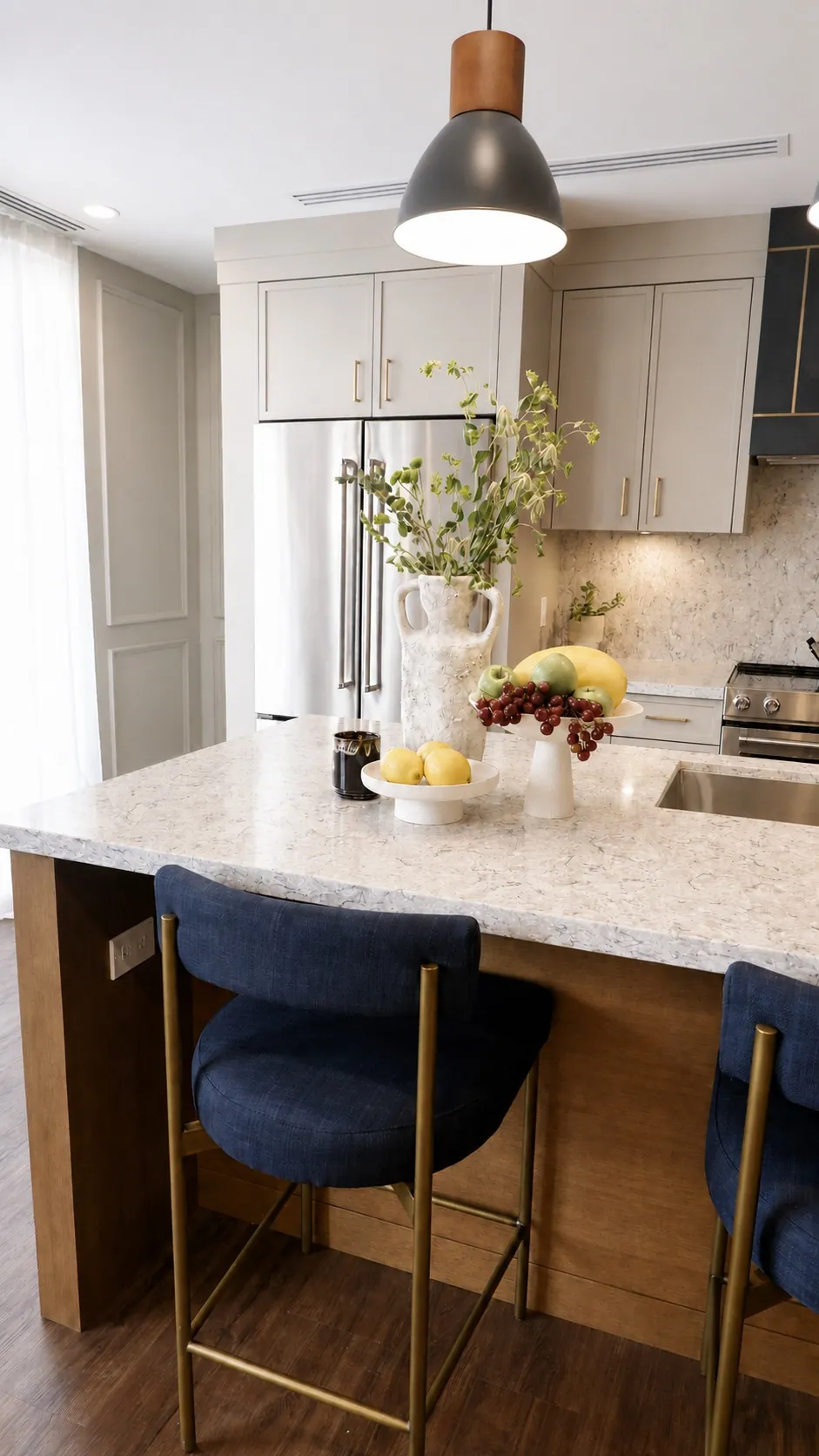 A modern kitchen features a marble island with two blue cushioned stools, a vase with greenery, a bowl of fruit, and pendant lighting. Light cabinetry and a stainless steel fridge are visible in the background.