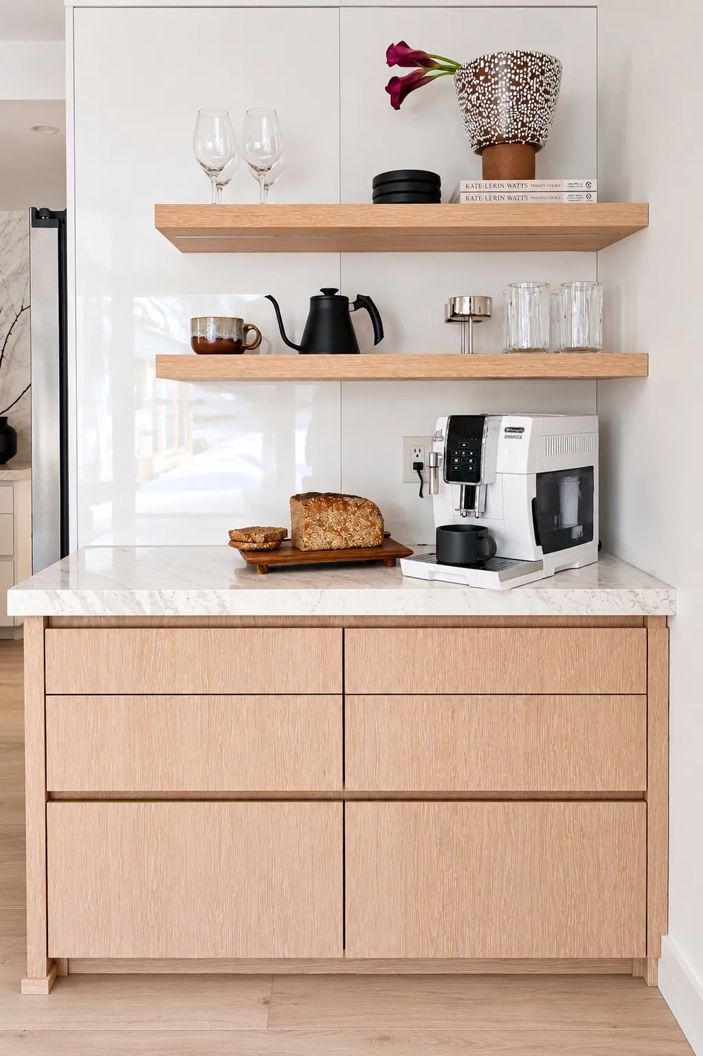 A modern kitchen corner with light wood cabinets, marble countertop, espresso machine, bread on a cutting board, two floating shelves with glasses, cups, a teapot, books, and a vase with dark pink flowers.
