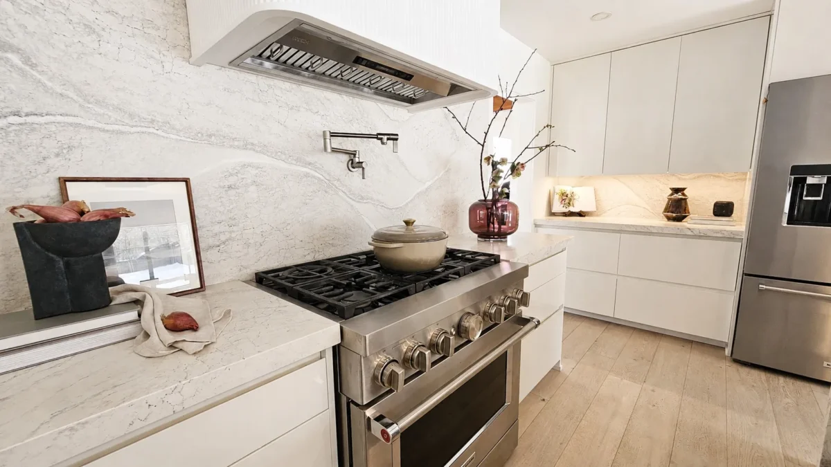 Modern kitchen with a stainless steel stove and pot, marble countertops and backsplash, built-in cabinets, a vase with branches, and a refrigerator. Light wood flooring and decorative items create a clean, elegant look.