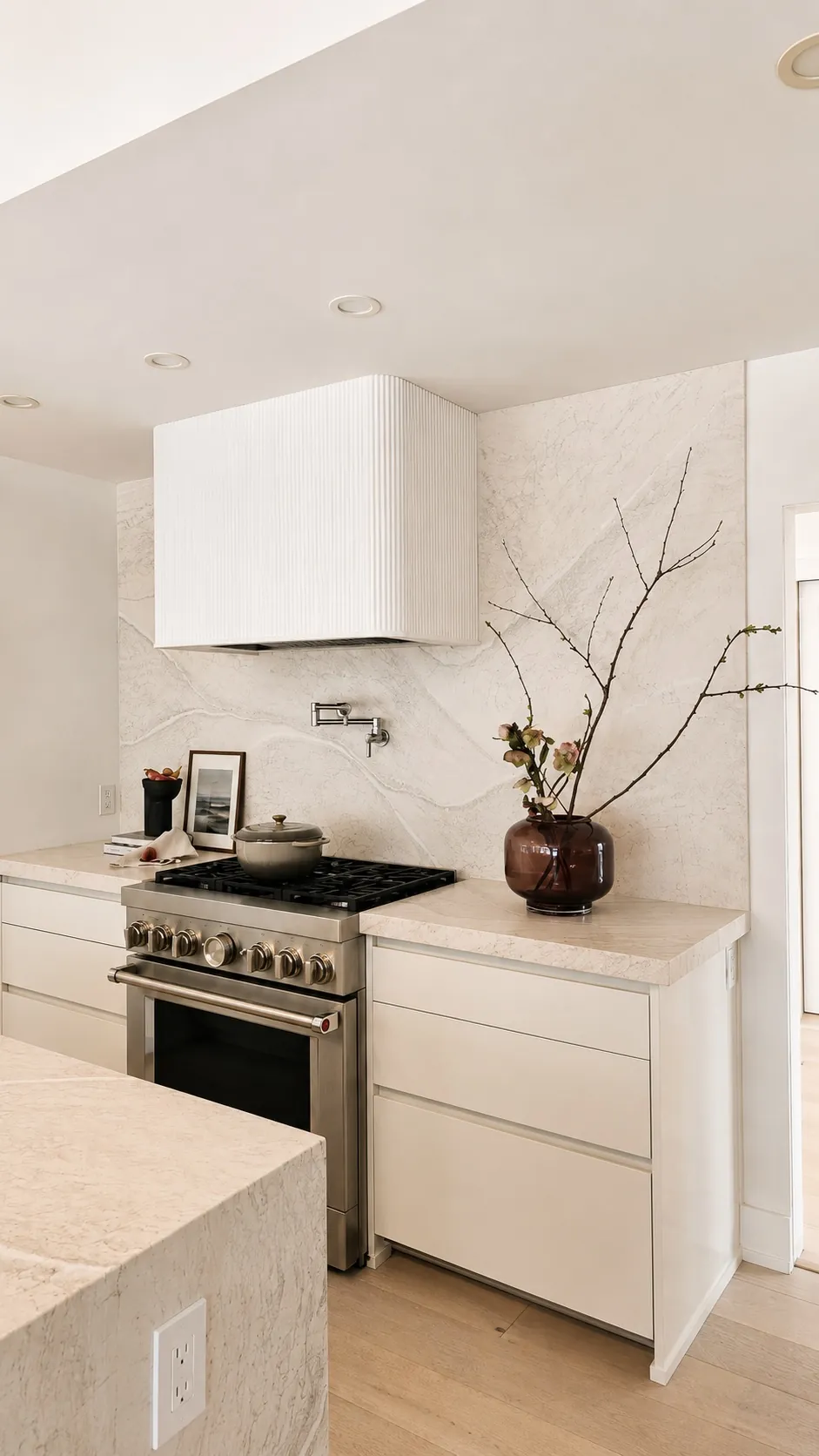 Minimalist kitchen with white cabinets, marble backsplash, stainless steel stove, a pot filler above the stove, and a brown vase with branches on the counter. Neutral tones and clean lines create a modern look.