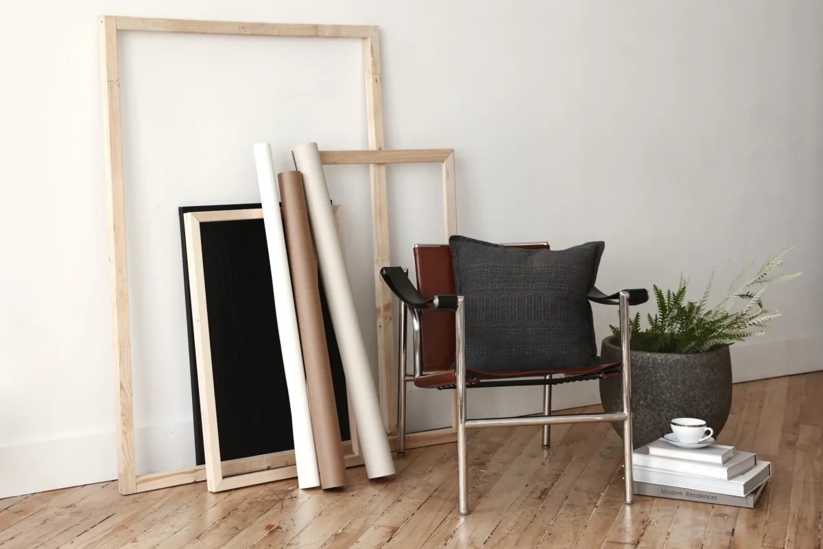 A minimalist room with wooden frames and rolled canvases leaning against a white wall, a modern chair with a dark cushion, a potted fern, and a stack of books with a cup and saucer on top, all on a wooden floor.