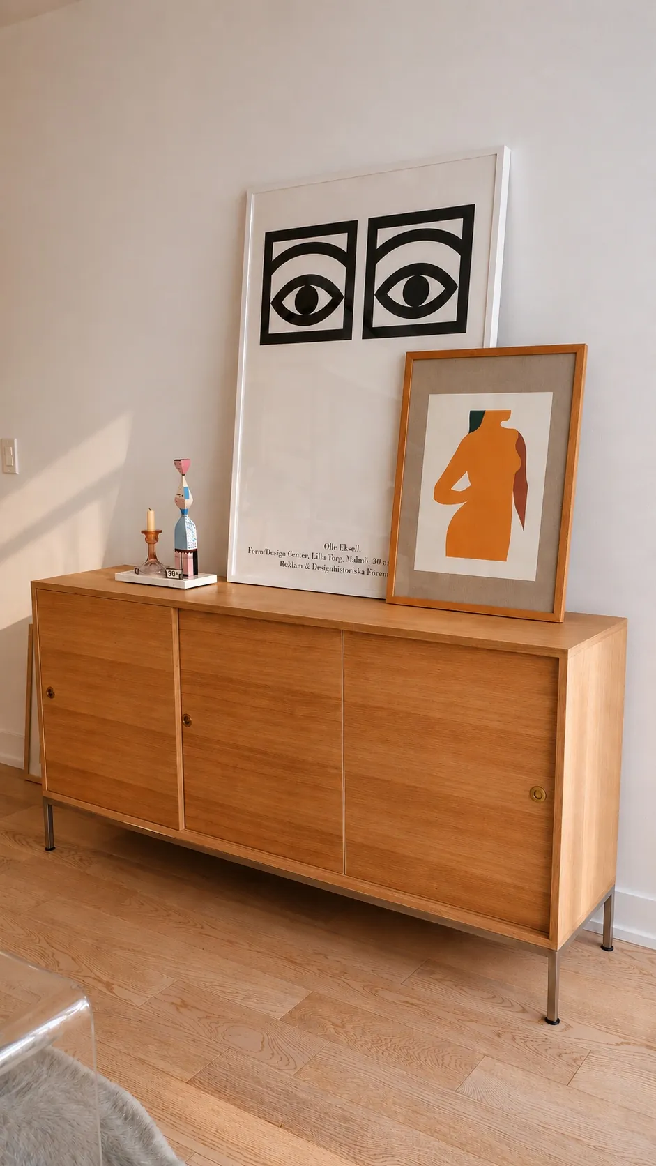 A wooden sideboard with metal legs stands against a white wall. On top are two framed artworks—one with bold black eyes, the other an abstract orange figure—along with a candleholder and a small decorative bottle.