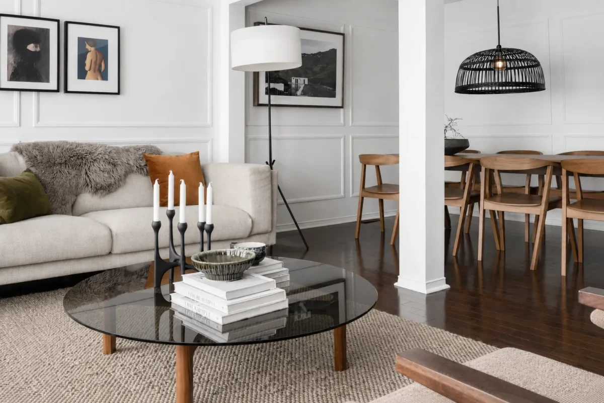 A modern living room with a beige sofa, fur throw, and green cushion, a glass coffee table with books and candles, and a dining area with wooden chairs, a black pendant lamp, and framed art on white paneled walls.