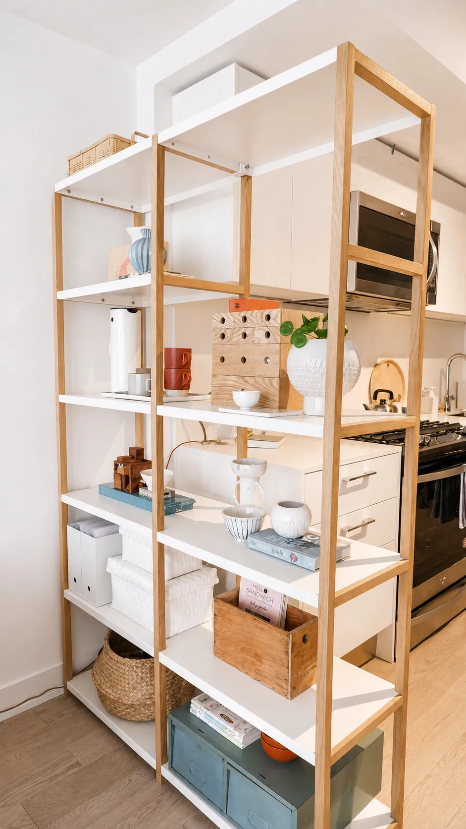 Open wooden shelving unit with white shelves, displaying books, baskets, ceramics, plants, and storage boxes. The shelf acts as a room divider between a modern kitchen and living space.