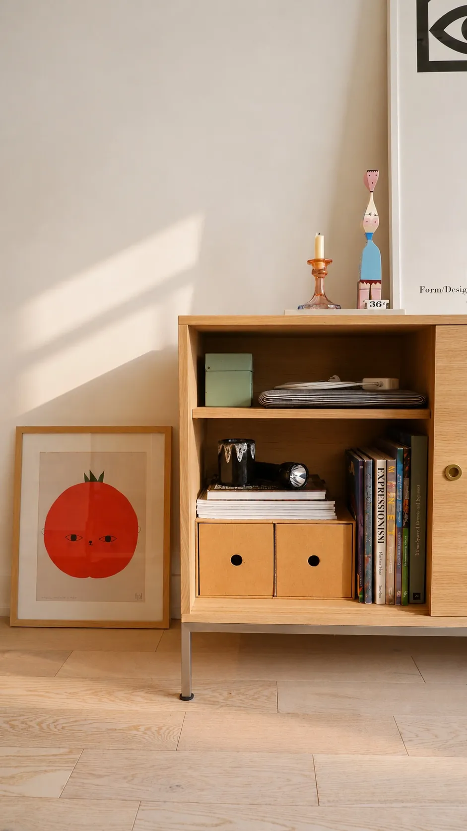 A light wood cabinet with books, boxes, a candle, and small figurines sits beside a framed tomato illustration on the floor, all against a light-colored wall in a sunlit room.