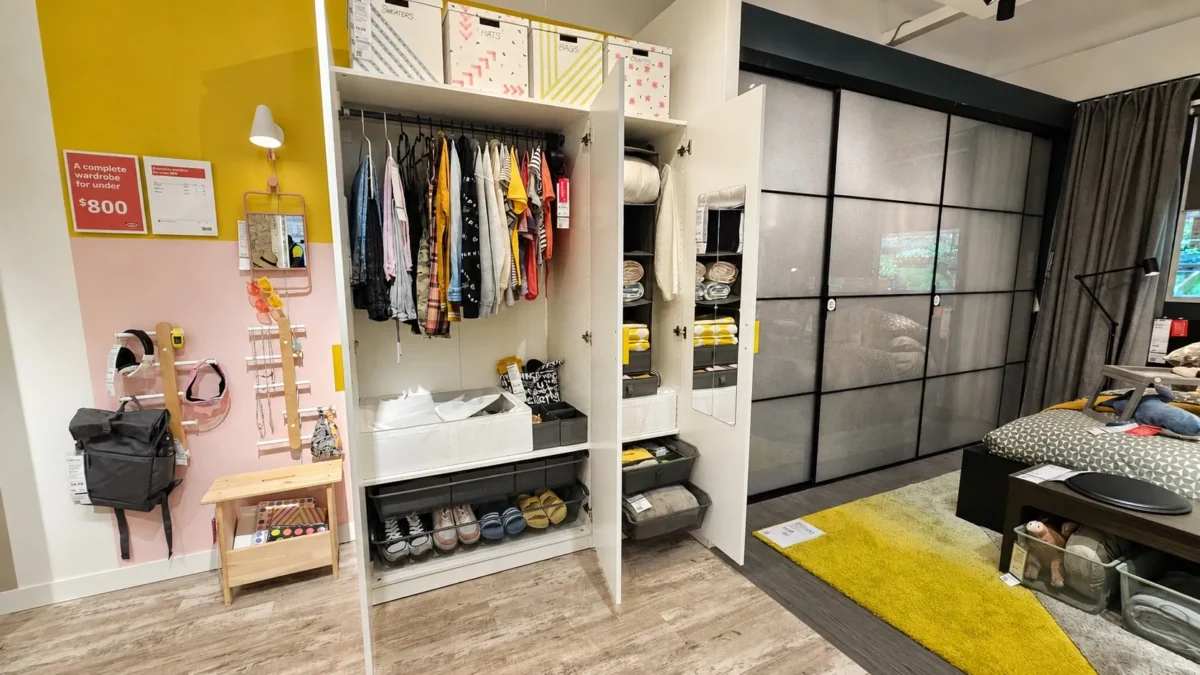 Open wardrobe with clothes, shoes, and storage boxes organized inside; a small wooden bench and hooks on a yellow wall to the left; a modern living space with a bed and glass sliding doors in the background.