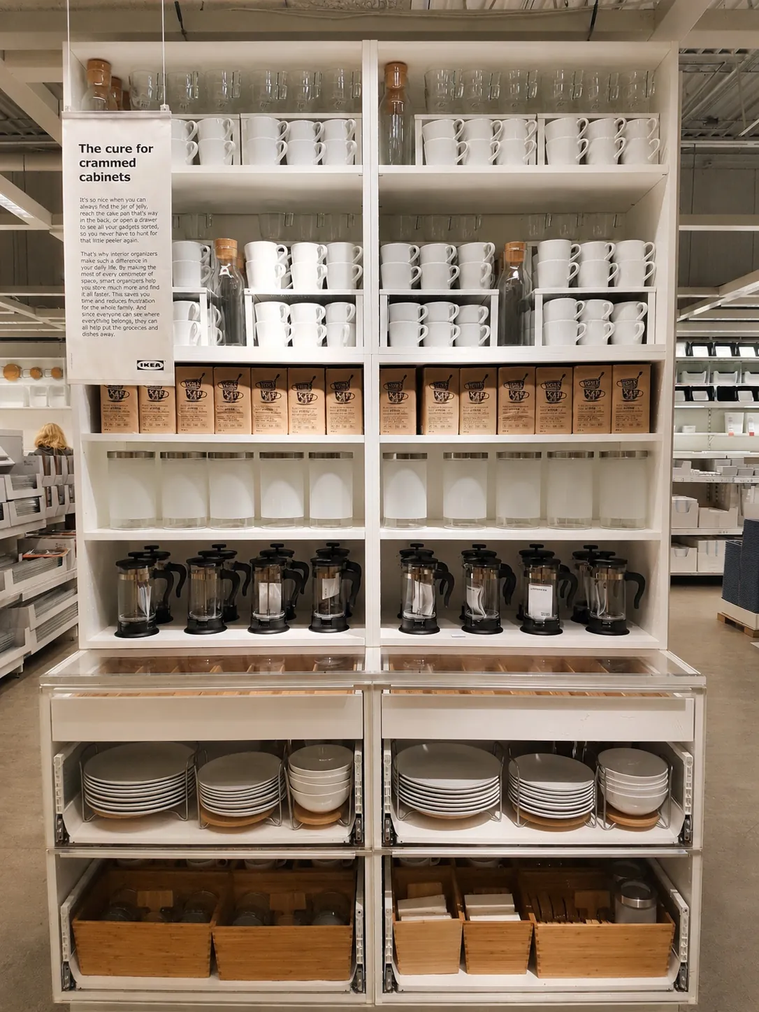A neatly organized store display features shelves with white mugs, glasses, jars, coffee makers, and boxes, with drawers below holding white dishes, bowls, and wooden trays. A sign with storage tips is on the upper left.