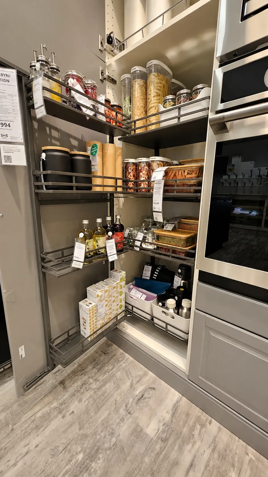 A modern kitchen pantry with pull-out shelves, neatly organized with jars of pasta, spices, cans, bottles, condiments, and black storage containers, next to a built-in oven and gray cabinetry.