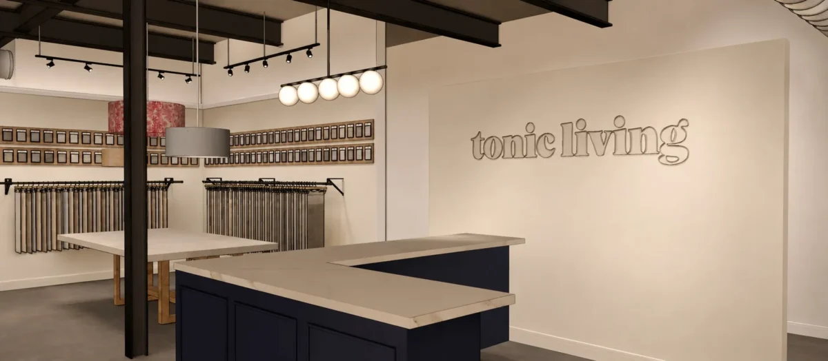 Modern retail store interior with a front counter, fabric sample displays, hanging lights, and brand signage on a cream-colored wall. Fabric rolls and sample swatches are organized along the walls and racks.