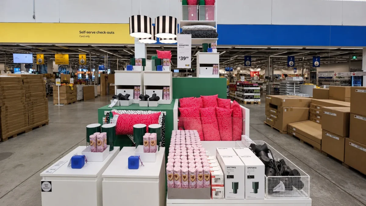 A display of home decor items including pink and black cushions, lamps, candles, and tumblers is set up in the middle of a large store with checkout counters and stacked boxes in the background.