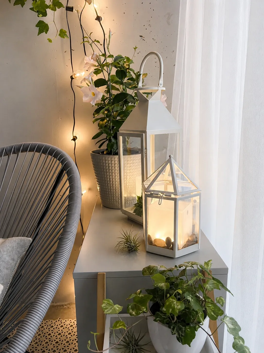 A cozy corner with potted plants, two white lanterns—one larger than the other—with candles inside, glowing softly. Twinkle lights hang on the wall beside a gray chair with a cushion and sheer white curtains.