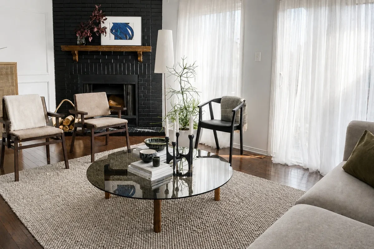 A modern living room with a glass coffee table, beige chairs, a light gray sofa, a textured rug, and a black brick fireplace. Sheer curtains cover tall windows, letting in soft natural light.