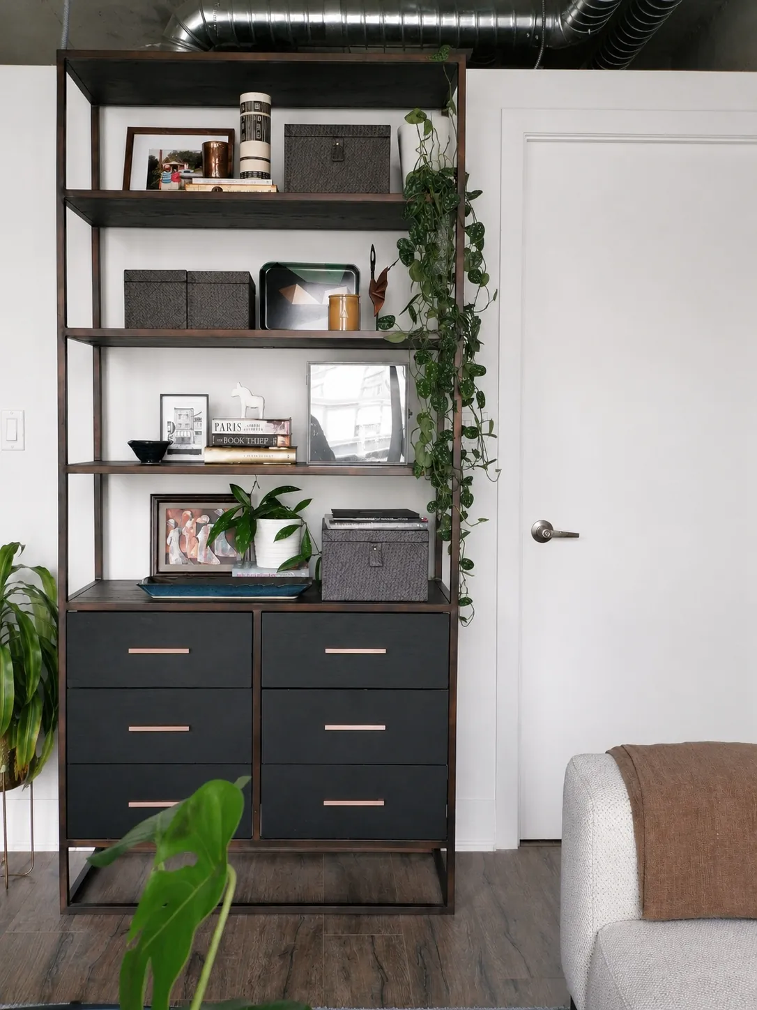 A tall black shelving unit with cabinets and drawers holds books, baskets, plants, candles, and decorative items. It stands against a white wall near a closed door and a gray armchair, with greenery in the foreground.