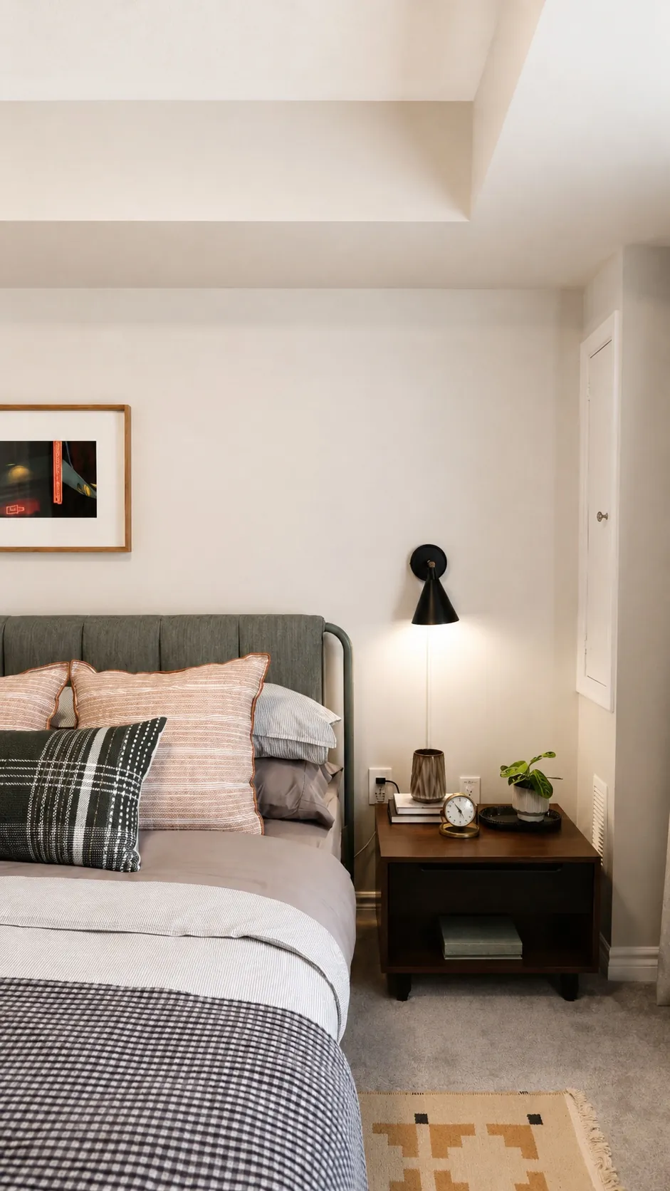 A neatly made bed with layered pillows and neutral bedding sits beside a dark wood nightstand holding a lamp, books, clock, and small plant. Framed art hangs above on a light-colored bedroom wall.