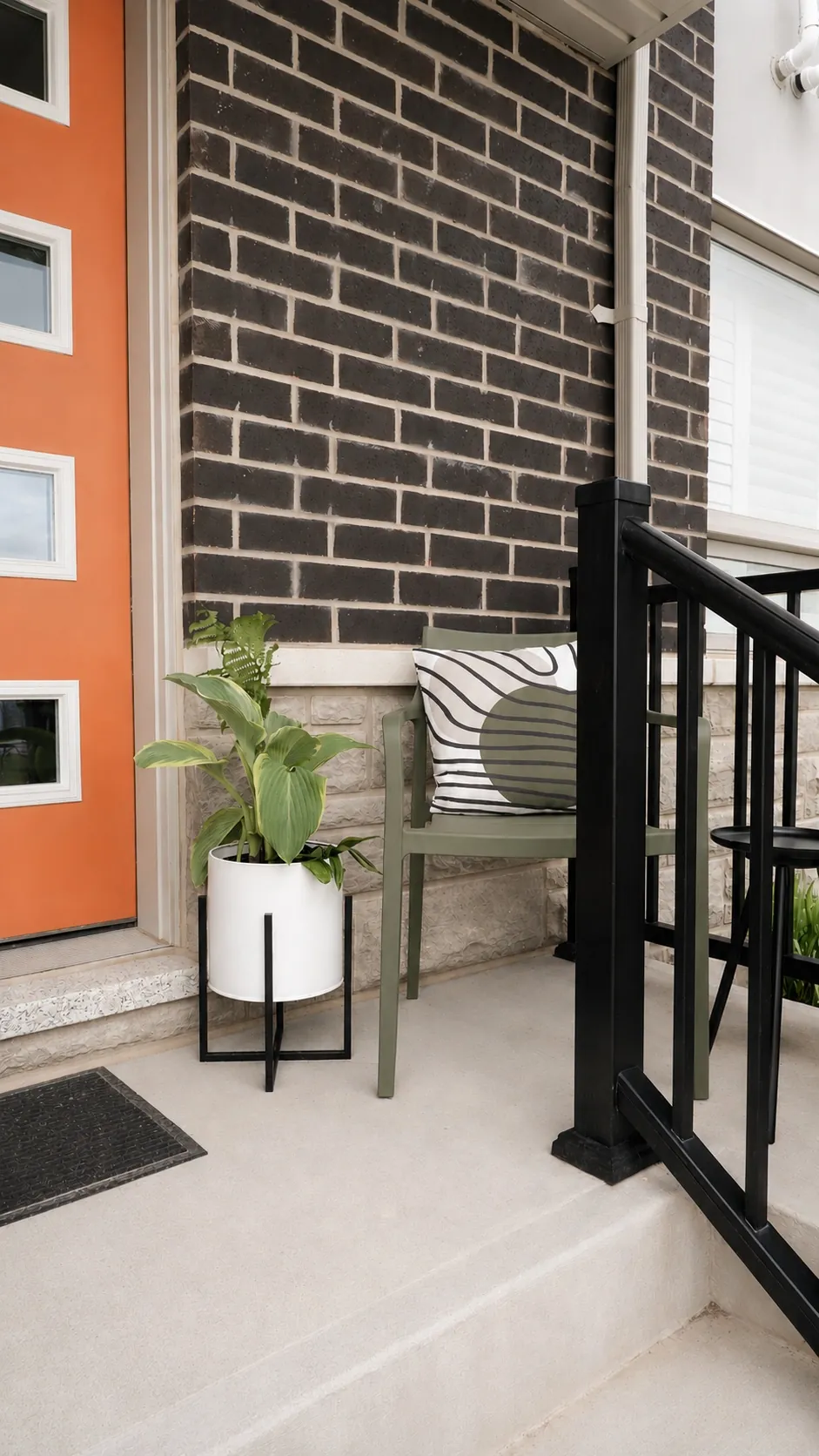 A modern porch with a bright orange door, black brick wall, olive green chair with a patterned cushion, a potted plant in a white planter, and a black metal railing.