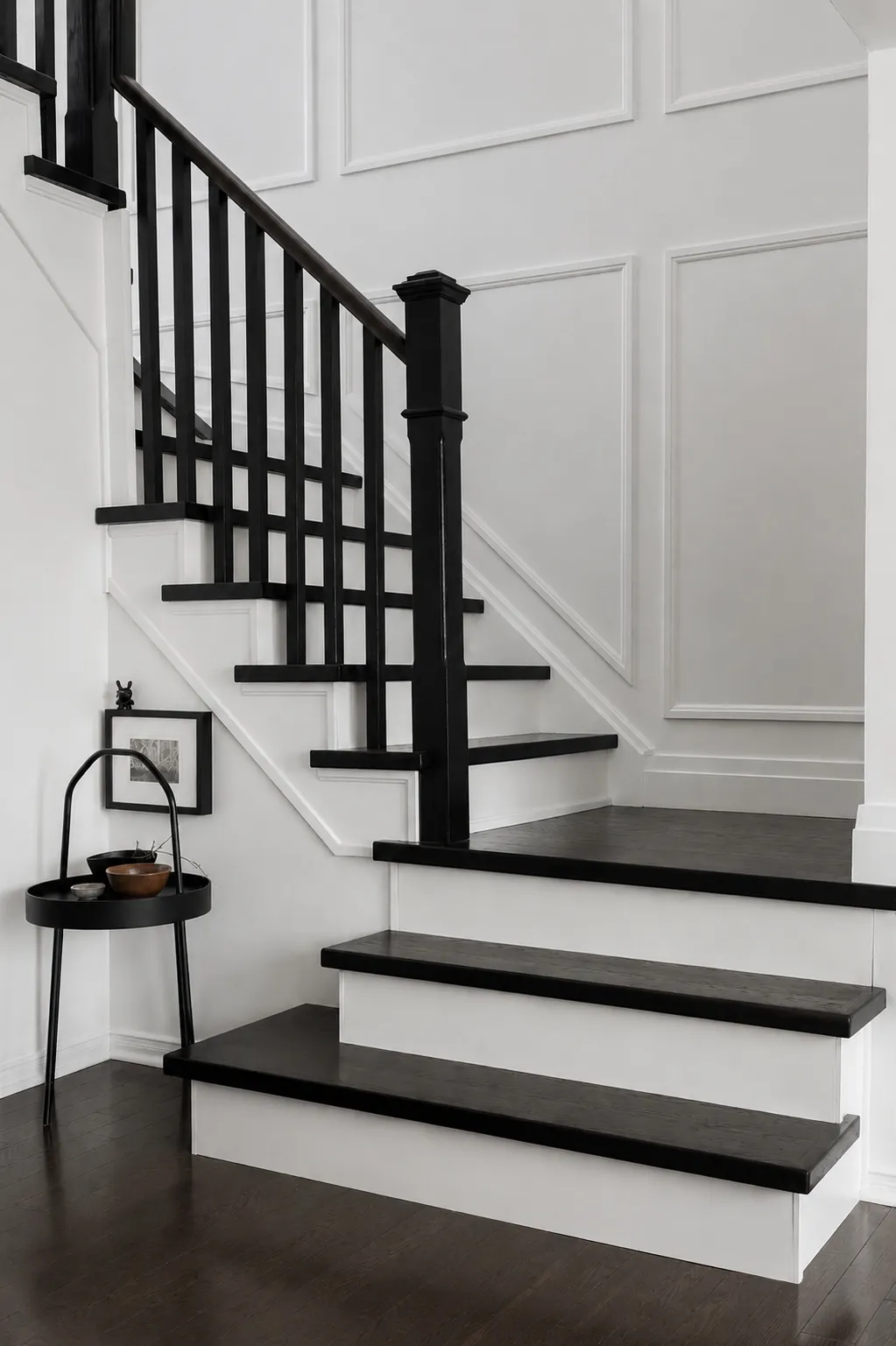 A modern staircase with black handrails and steps, set against white paneled walls. A small black side table with decor sits at the base of the stairs on dark wood flooring.