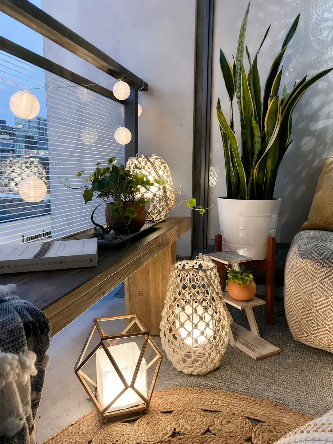 A cozy balcony with string lights, potted plants, and glowing lanterns on a woven rug; a wooden bench holds books, a knitted blanket, and more plants, creating a warm, inviting atmosphere at dusk.