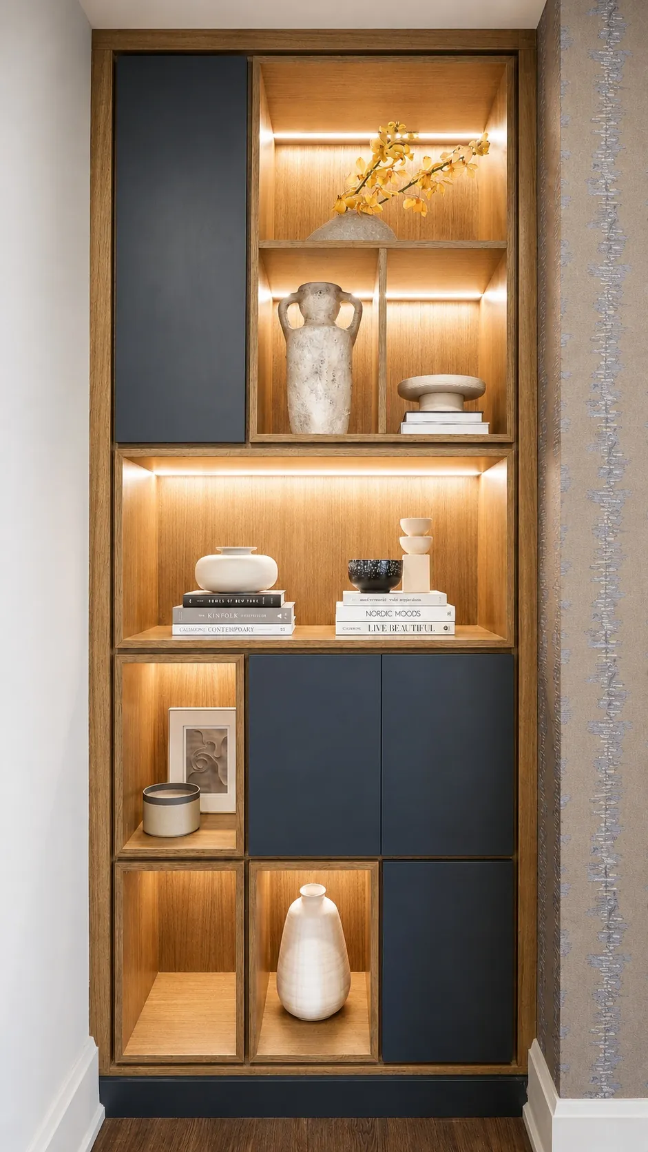 A built-in wooden shelving unit with black cabinet doors, featuring books, vases, bowls, a framed photo, and decorative objects, all softly lit by recessed lighting.