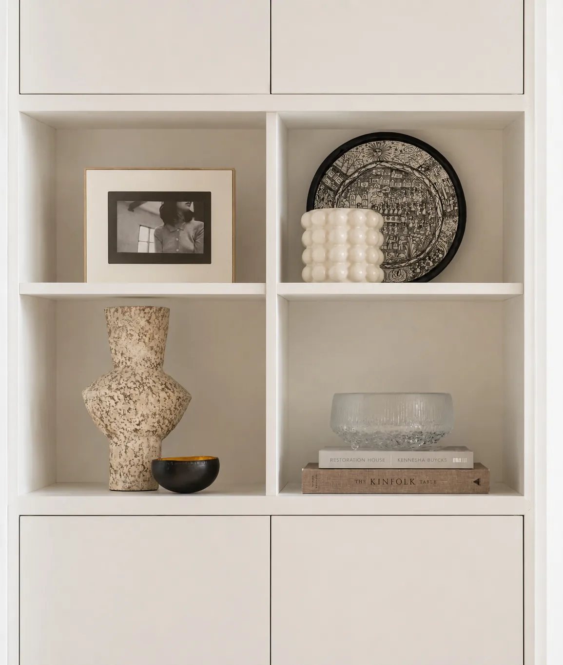 A minimalist shelf displays a framed photo, a decorative plate, a white bubble candle, a speckled vase, a small bowl, a glass bowl, and stacked books, all arranged neatly on light-colored shelves.