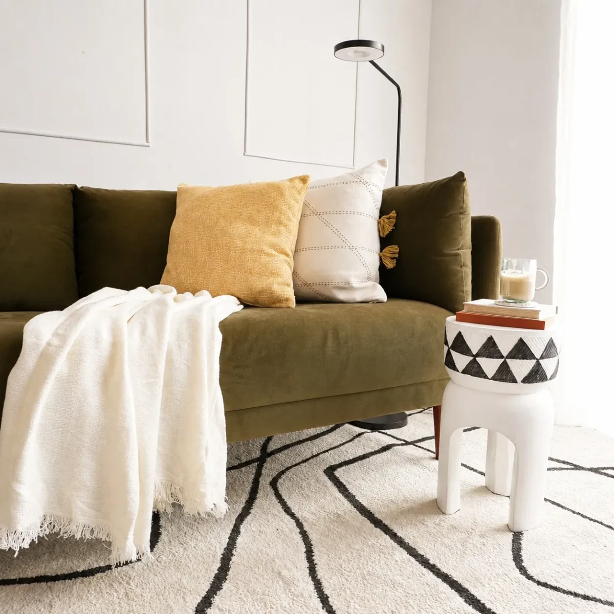 A modern living room with an olive green sofa, two pillows, a white throw blanket, a small white side table with a geometric pattern, a glass of coffee, a rug with black lines, and a black floor lamp.
