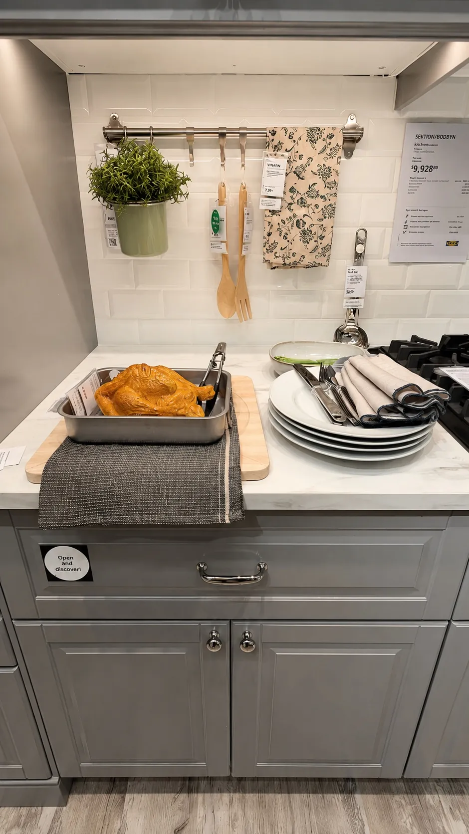 A modern kitchen counter with a baking tray holding a cooked chicken, stacked plates with rolled napkins, wooden utensils, a hanging potted plant, and a floral oven mitt against a white tile backsplash.