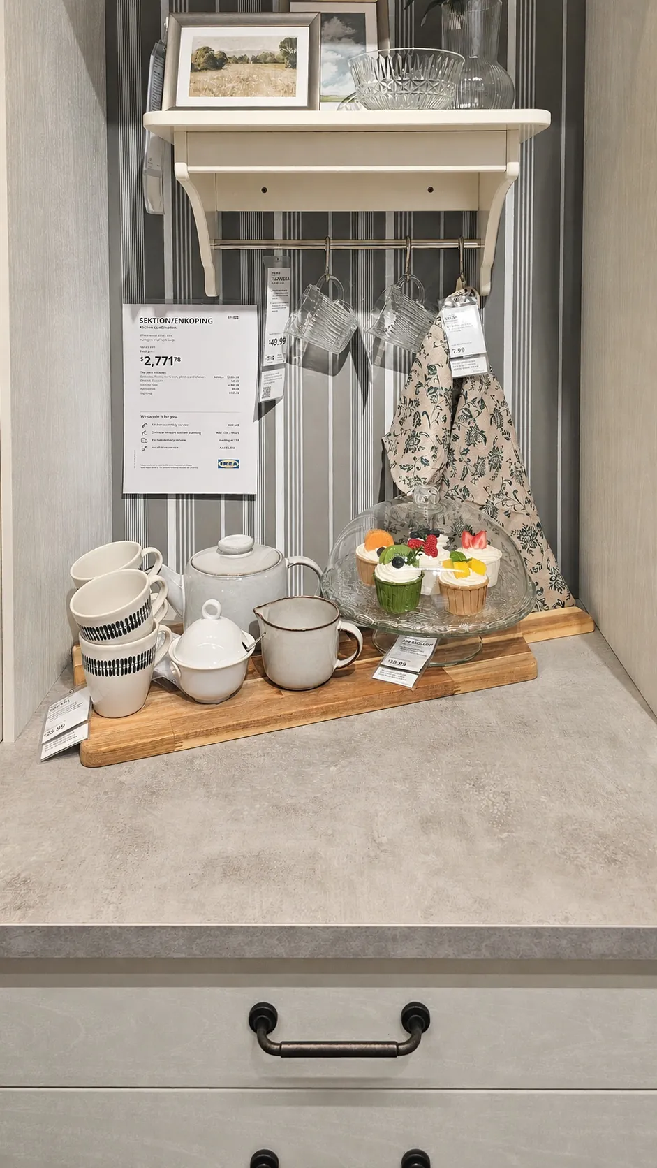 A kitchen display with a gray countertop, a wooden tray holding teacups, a teapot, a mug, and desserts. On the wall, there are glasses hanging and floral-patterned towels under a shelf with decor and price tags.