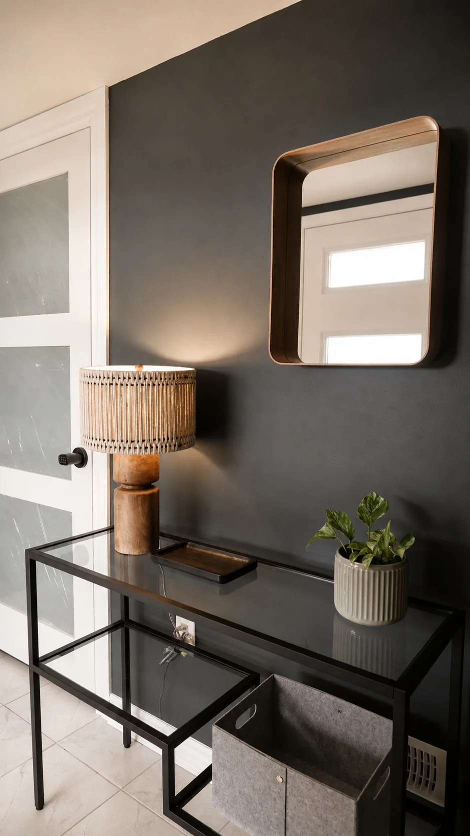 A modern entryway with a black console table, a rattan table lamp, a potted plant, a square mirror on a dark wall, and a gray storage bin on the lower shelf. A white door with frosted glass panels is nearby.