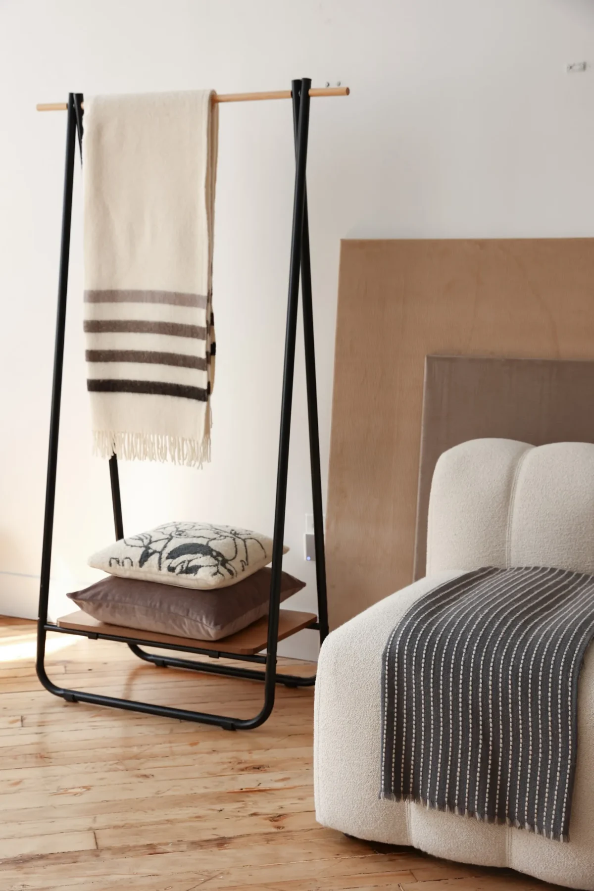 A cozy living space with a white boucle chair draped with a striped gray blanket, next to a black metal clothes rack holding a cream throw and stacked cushions on a wooden floor.