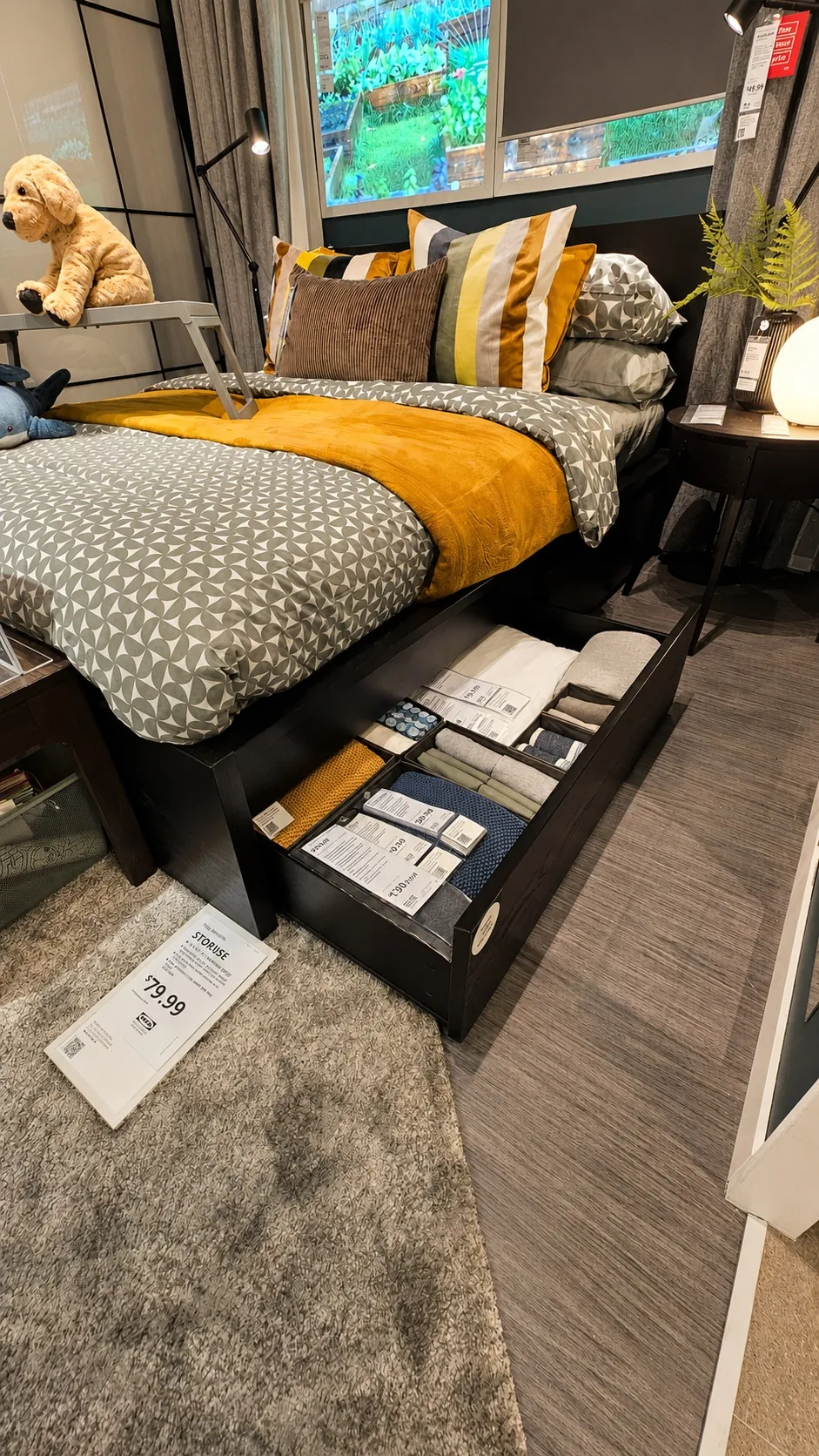 A neatly made bed with gray and yellow bedding, decorative pillows, and a mustard blanket; below, an open drawer stores folded linens. A plush dog and bedside table are nearby in a cozy, modern bedroom.