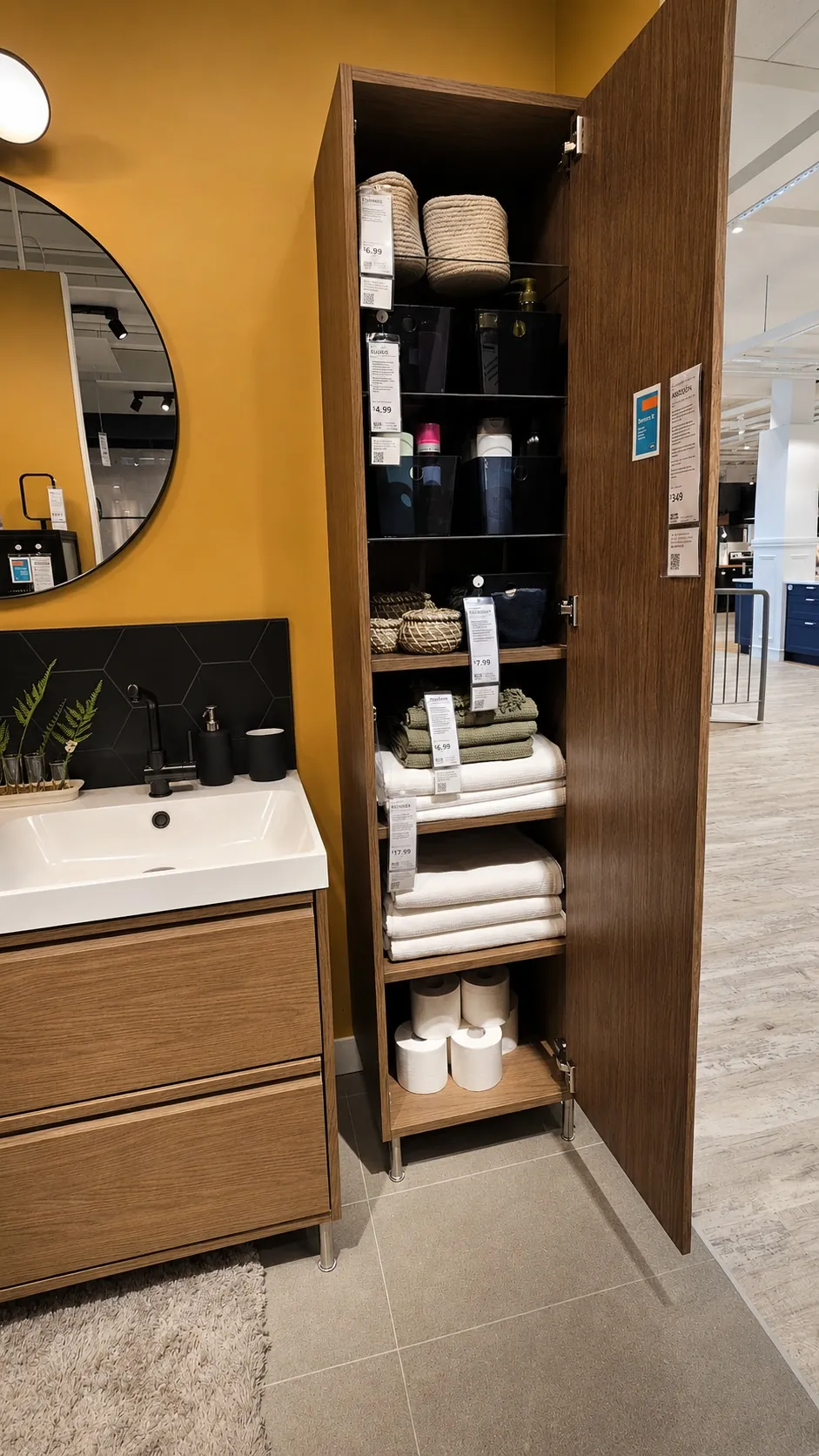 Tall wooden bathroom cabinet with open door revealing neatly organized towels, toilet paper, baskets, and toiletries. Next to it is a sink with a round mirror and yellow wall in a modern showroom setting.