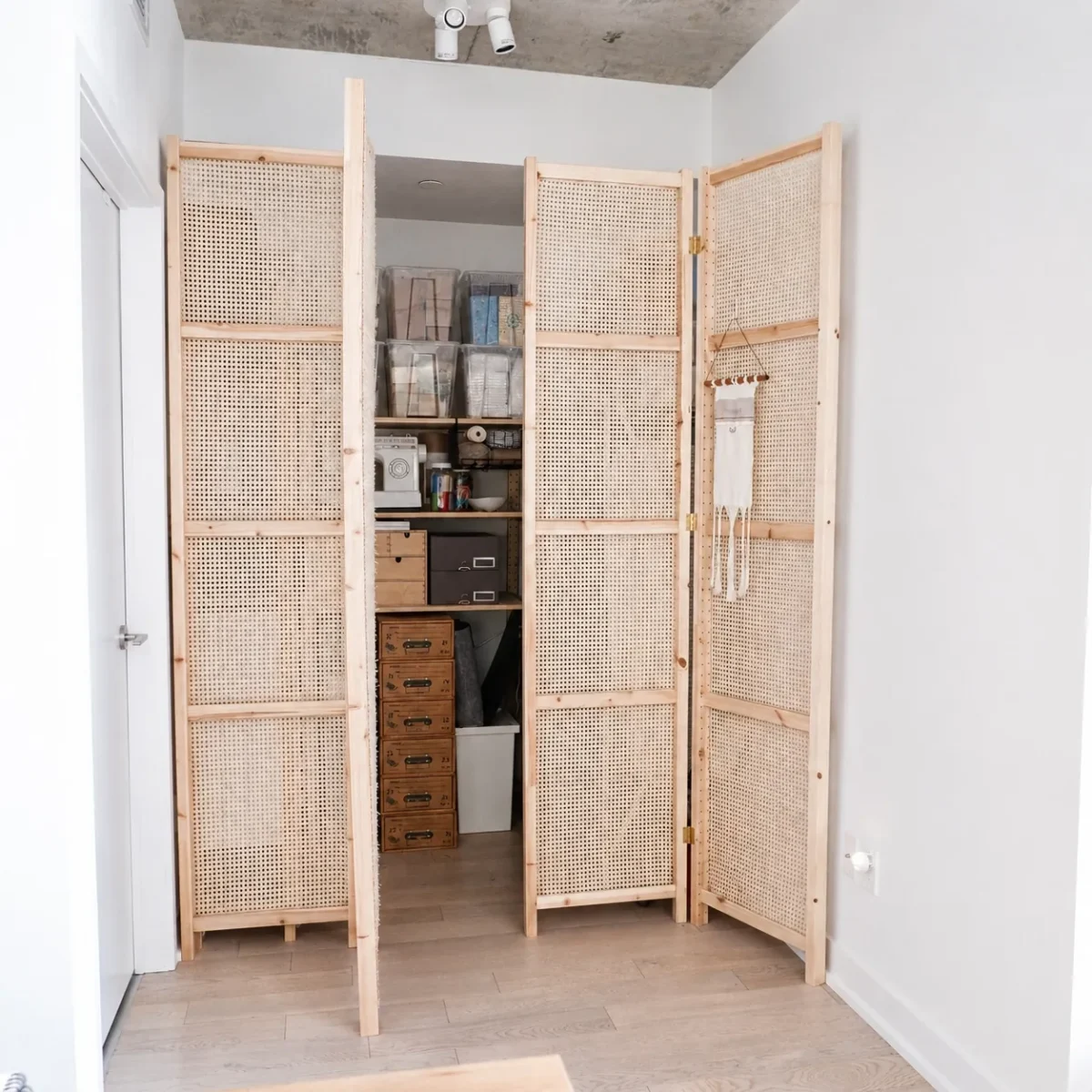 A light wooden folding room divider with woven panels partially conceals a closet filled with organized storage bins, boxes, and craft supplies in a bright, minimalist room with light wood flooring.