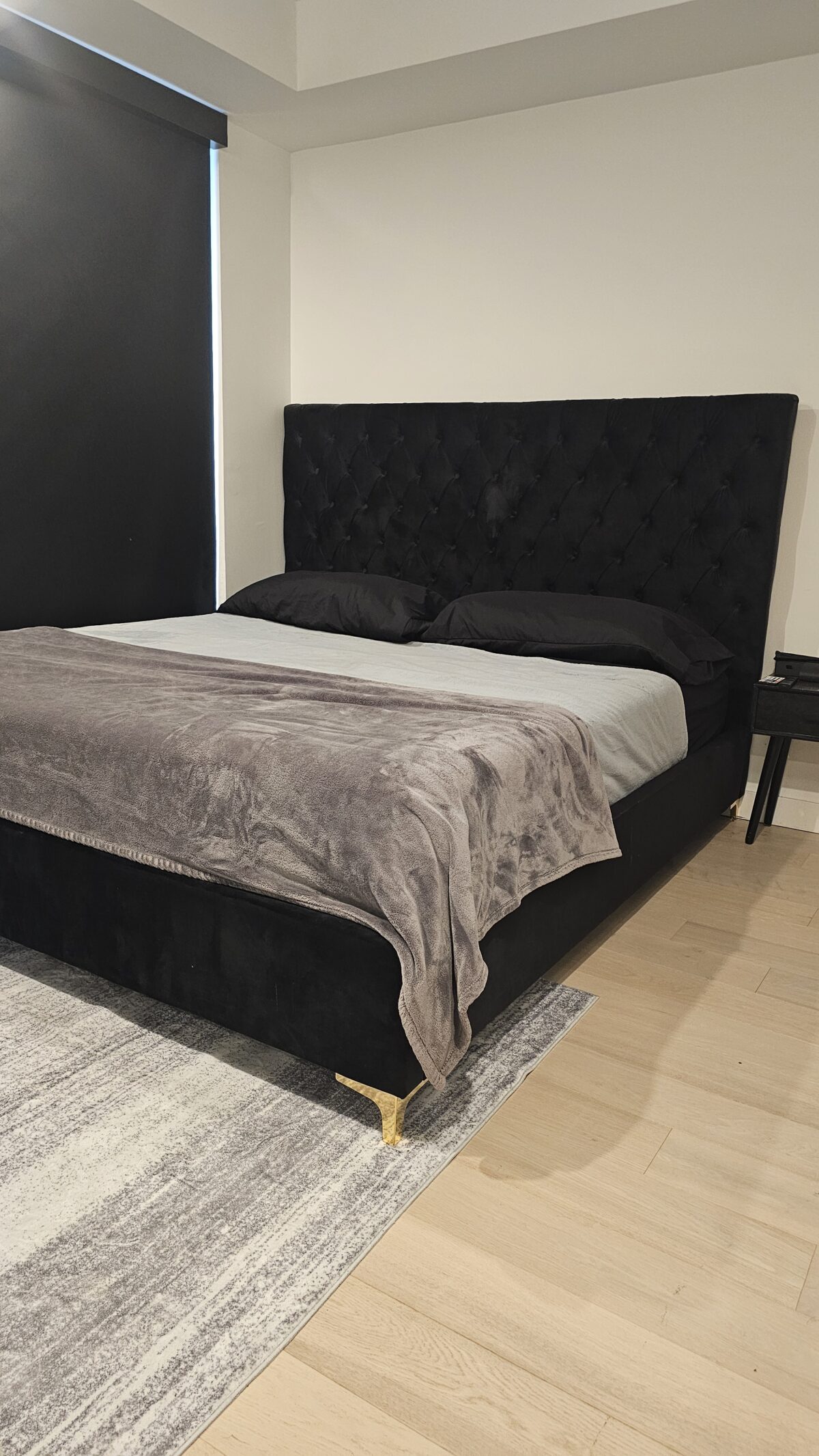 A modern bedroom with a black tufted headboard, black bedding, and a gray blanket on a large bed. There is a black nightstand beside the bed, a gray and white rug, and light wood floors. A window with a black blind is visible.