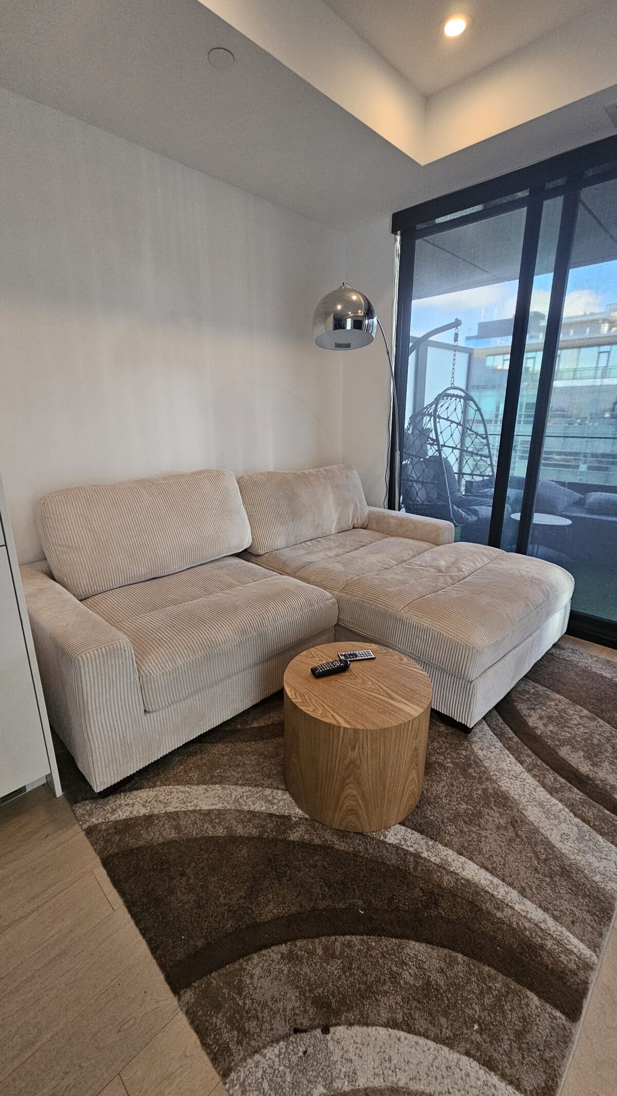 A modern living room with a light-colored sectional sofa, a round wooden coffee table, a floor lamp, and a large glass sliding door leading to a balcony with a hanging chair and urban view.