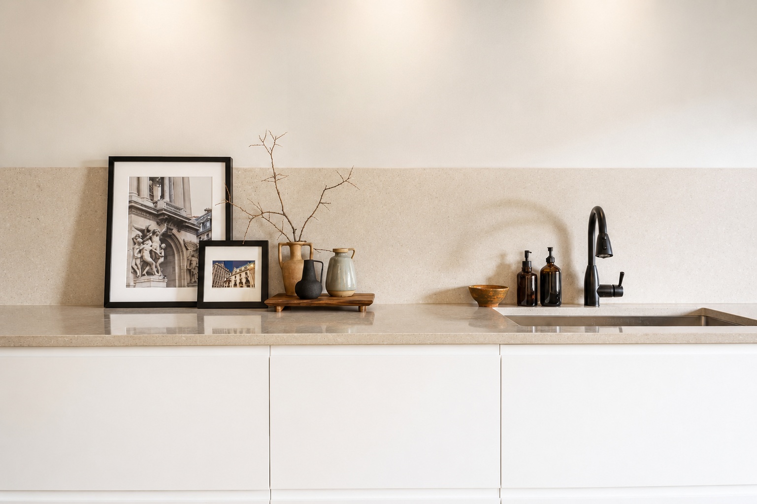Minimalist kitchen with a beige countertop, framed art, vases, a branch, soap bottles, and a black faucet. The background wall is light with soft overhead lighting.