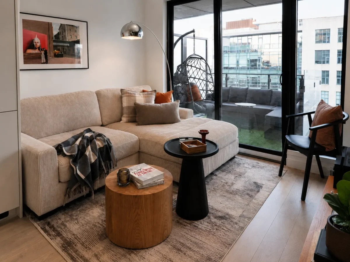 A modern living room with a beige sectional sofa, throw pillows, and a plaid blanket, two round coffee tables, a floor lamp, art on the wall, and large glass doors opening to a balcony with patio furniture and city views.