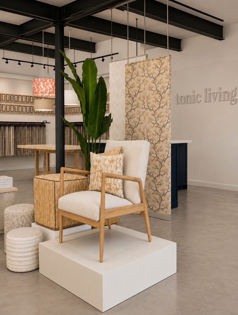 Contemporary showroom with a light wooden armchair on a white platform, patterned cushions, a tall green plant, textured cubes, and various fabric samples displayed in the background under bright lighting.