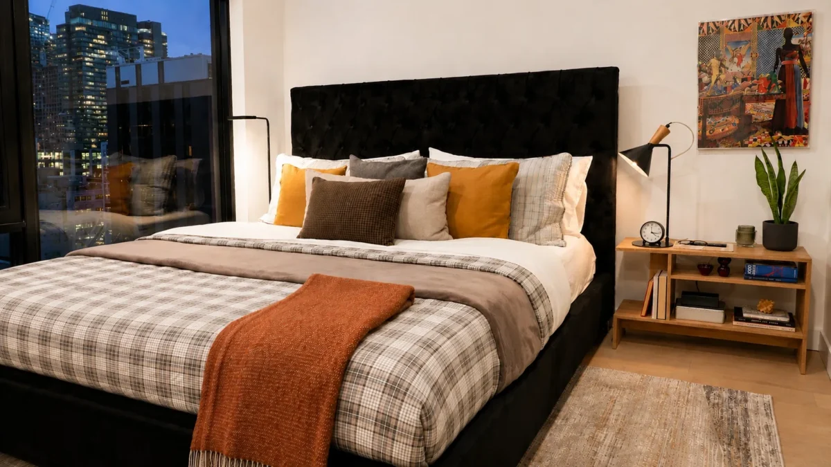 A modern bedroom with a large bed featuring plaid and neutral bedding, colorful accent pillows, and an orange throw blanket. There’s a window with a city view, two floor lamps, a wood nightstand, and a framed artwork on the wall.