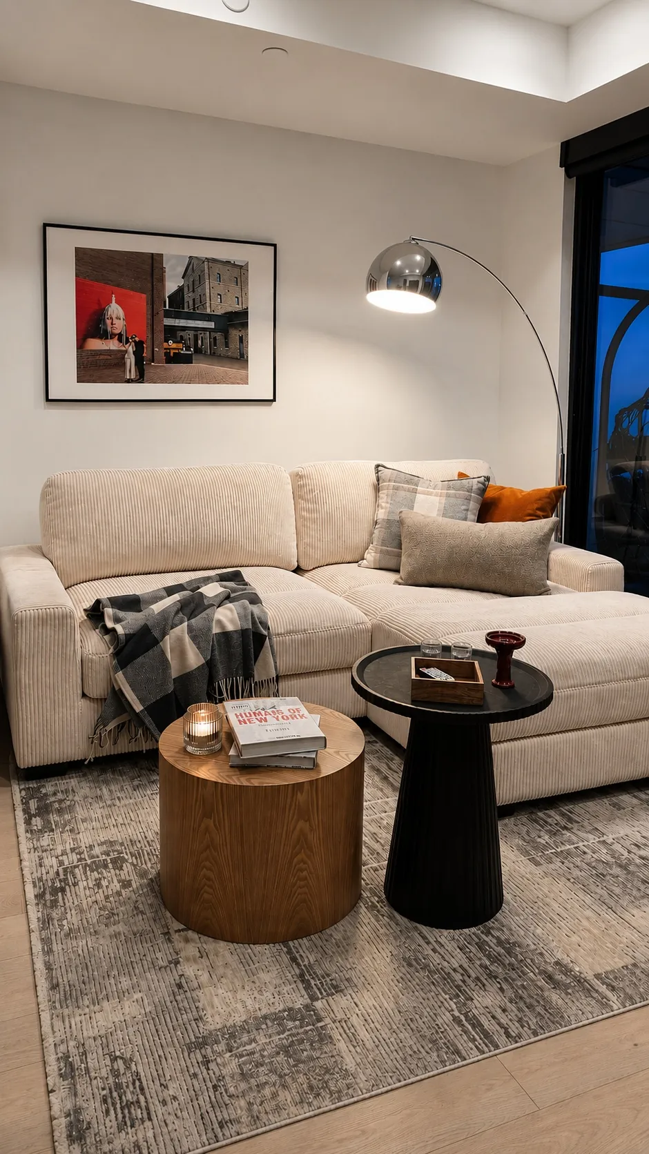 Modern living room with a beige sectional sofa, patterned throw blanket, pillows, two round coffee tables with books and decor, a floor lamp, and framed artwork on the wall above the sofa.